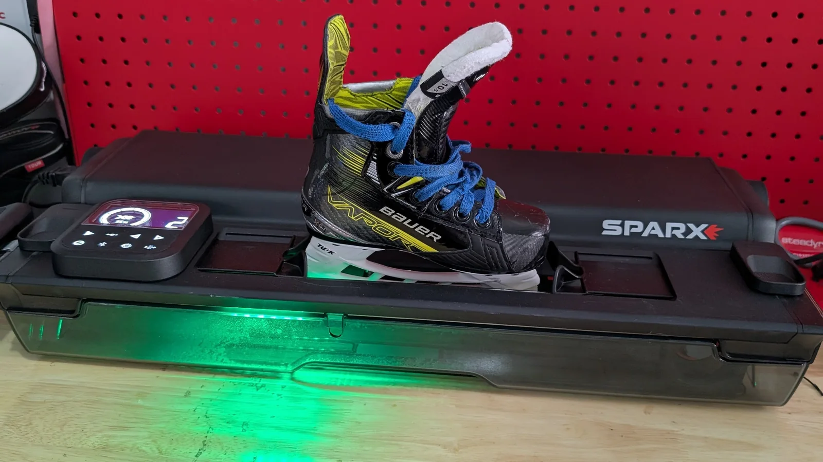 A Bauer skate resting on the SPARX sharpener, green light glowing, against a red pegboard